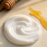 Closeup view of Milk and Honey Body Lotion next to smear of honey with a honey stick in the background