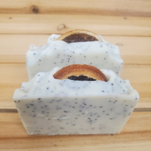 Two bars of Lemon poppy seed vegan soap with orange slices on a wooden surface
