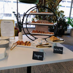 Three-tiered dessert stand with various pastries on a table in a room with large windows.